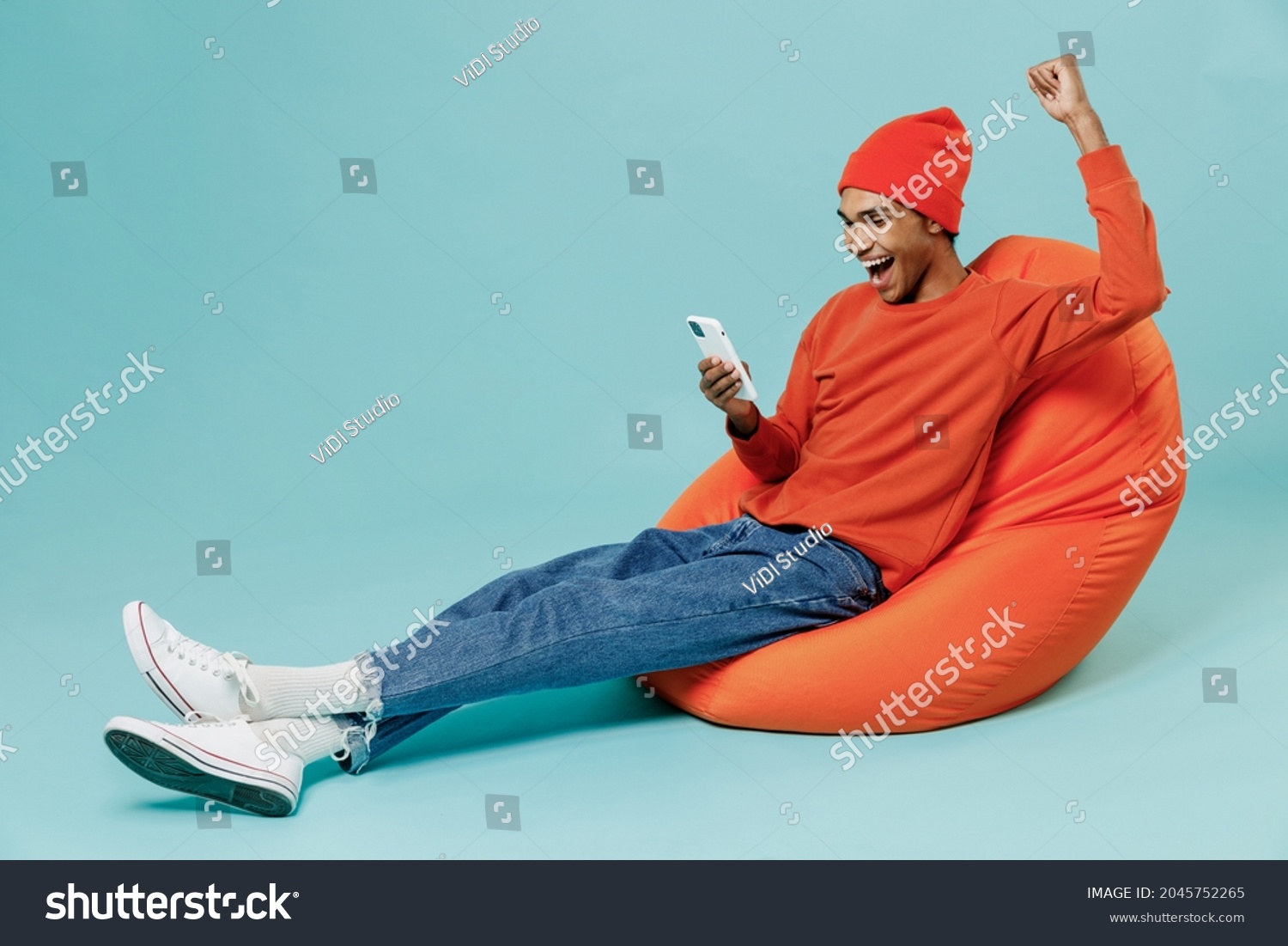 Full body young overjoye fun happy african american man wearing orange shirt hat sit in bag chair use hold mobile cell phone do winner gesture isolated on plain pastel light blue background studio. #2045752265