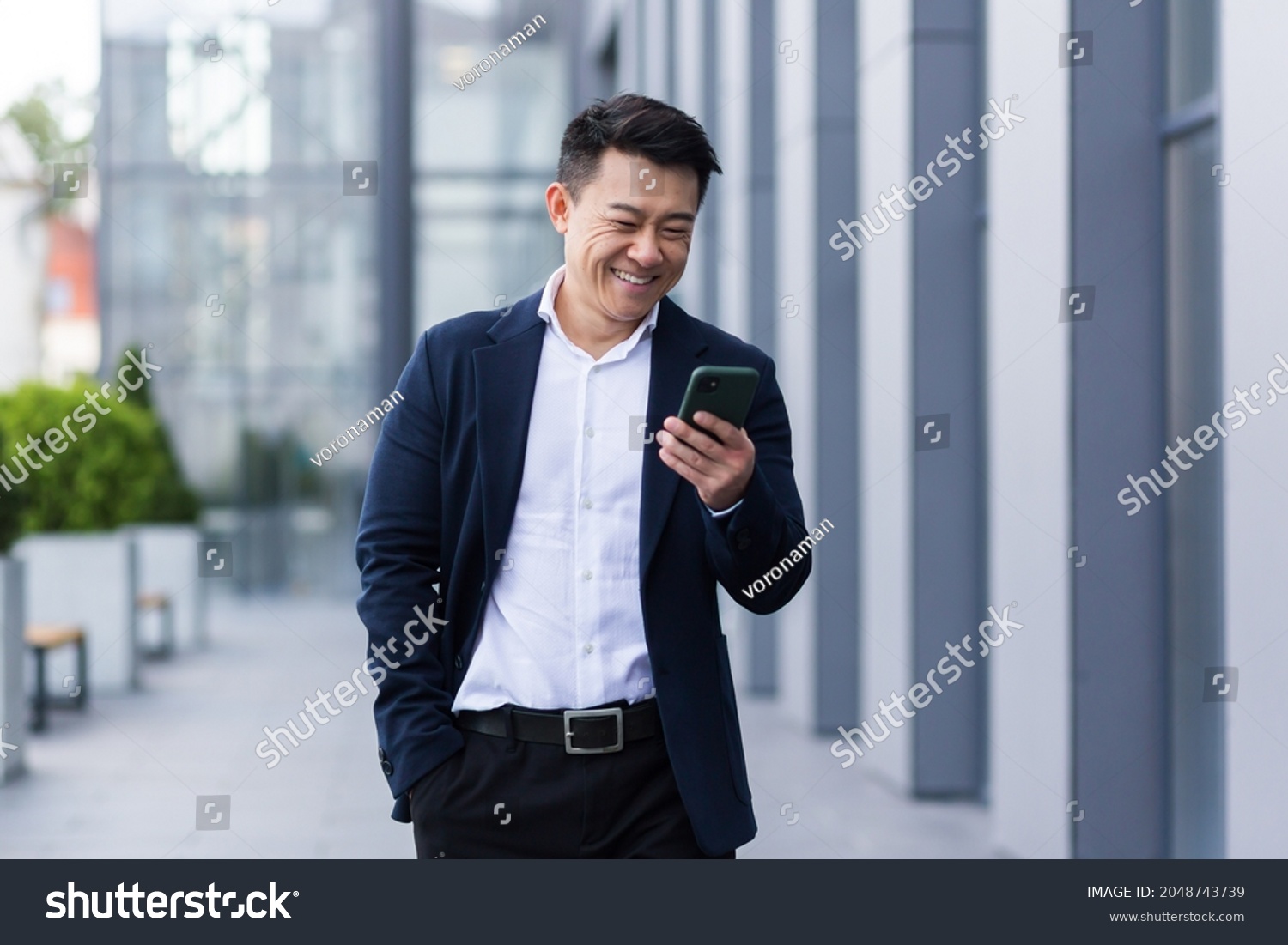 asian male freelancer walking near business center holding phone, smiling reading news, successful businessman #2048743739