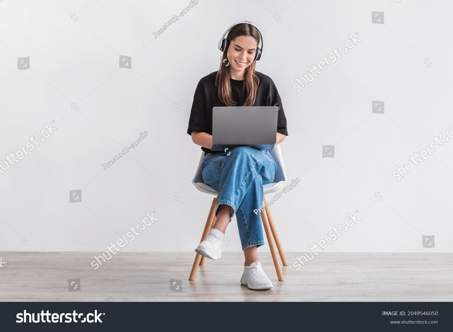 Cheery Caucasian woman in headphones having online video call on laptop, sitting on chair against white wall, full length. Millennial lady participating in webinar, communicating remotely on internet #2049546050