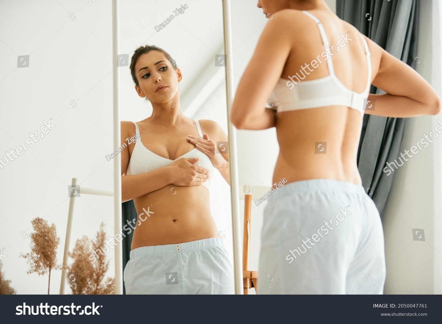 Young woman doing breast self-examination while looking herself in a mirror at home. #2050047761