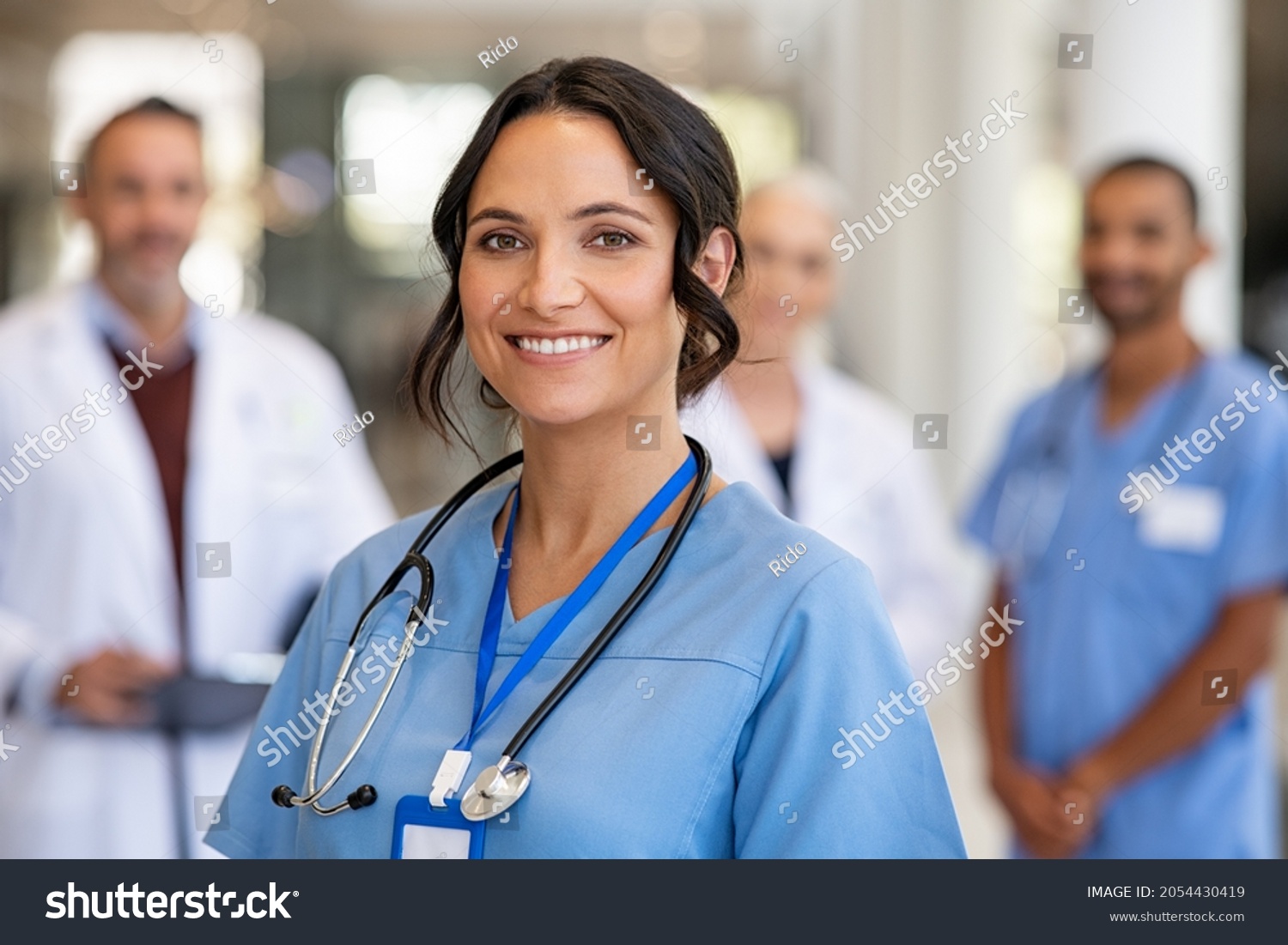 Portrait of happy young nurse in uniform with healthcare team in background. Successful team of doctor and nurses smiling. Beautiful and satisfied healthcare worker in private clinic looking at camera #2054430419
