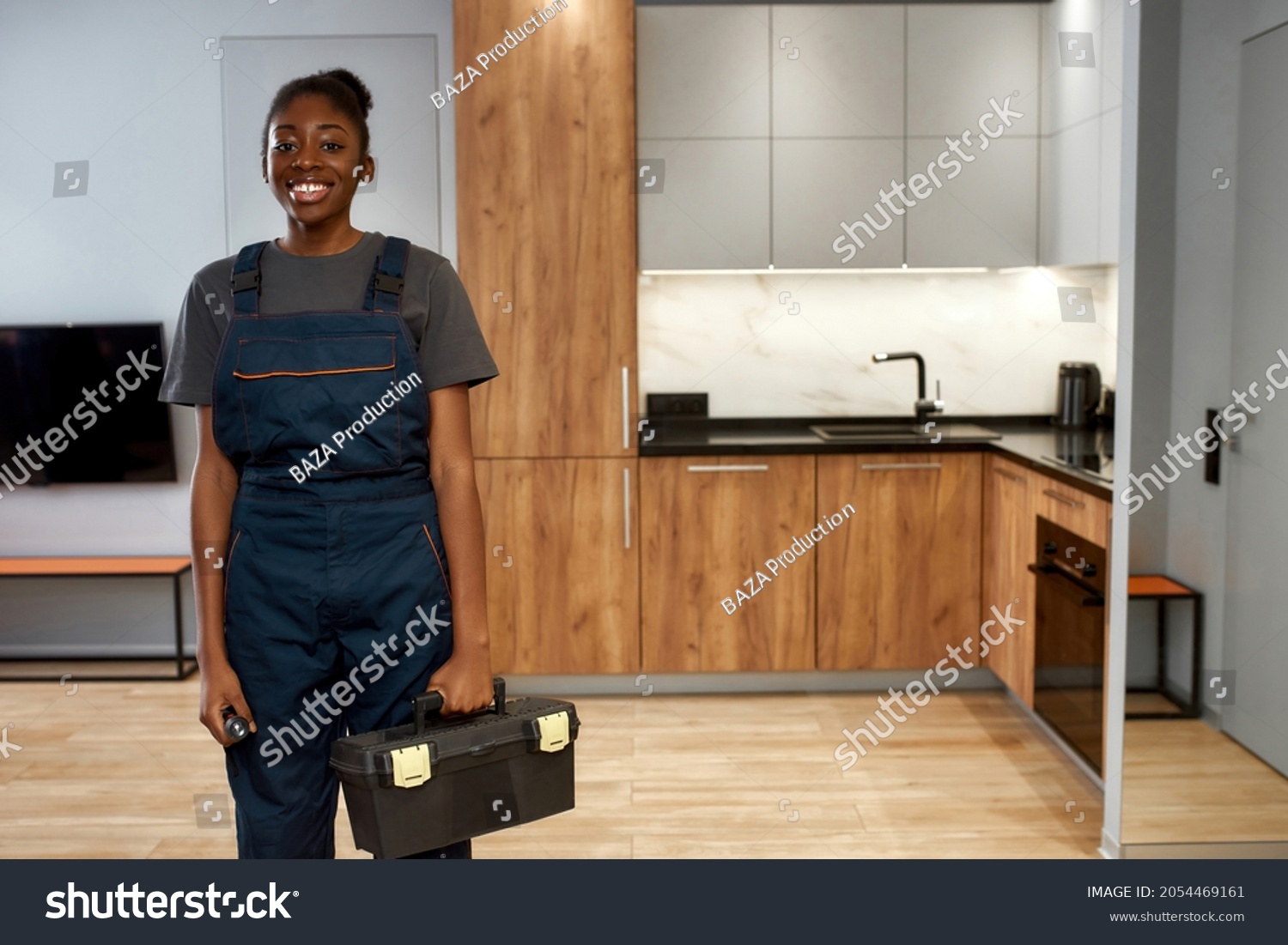 Indoor portrait of young smiling african american woman, professional technician or repairwoman posing in kitchen, holding toolkit. Female home repair service worker. Modern kitchen on background. #2054469161