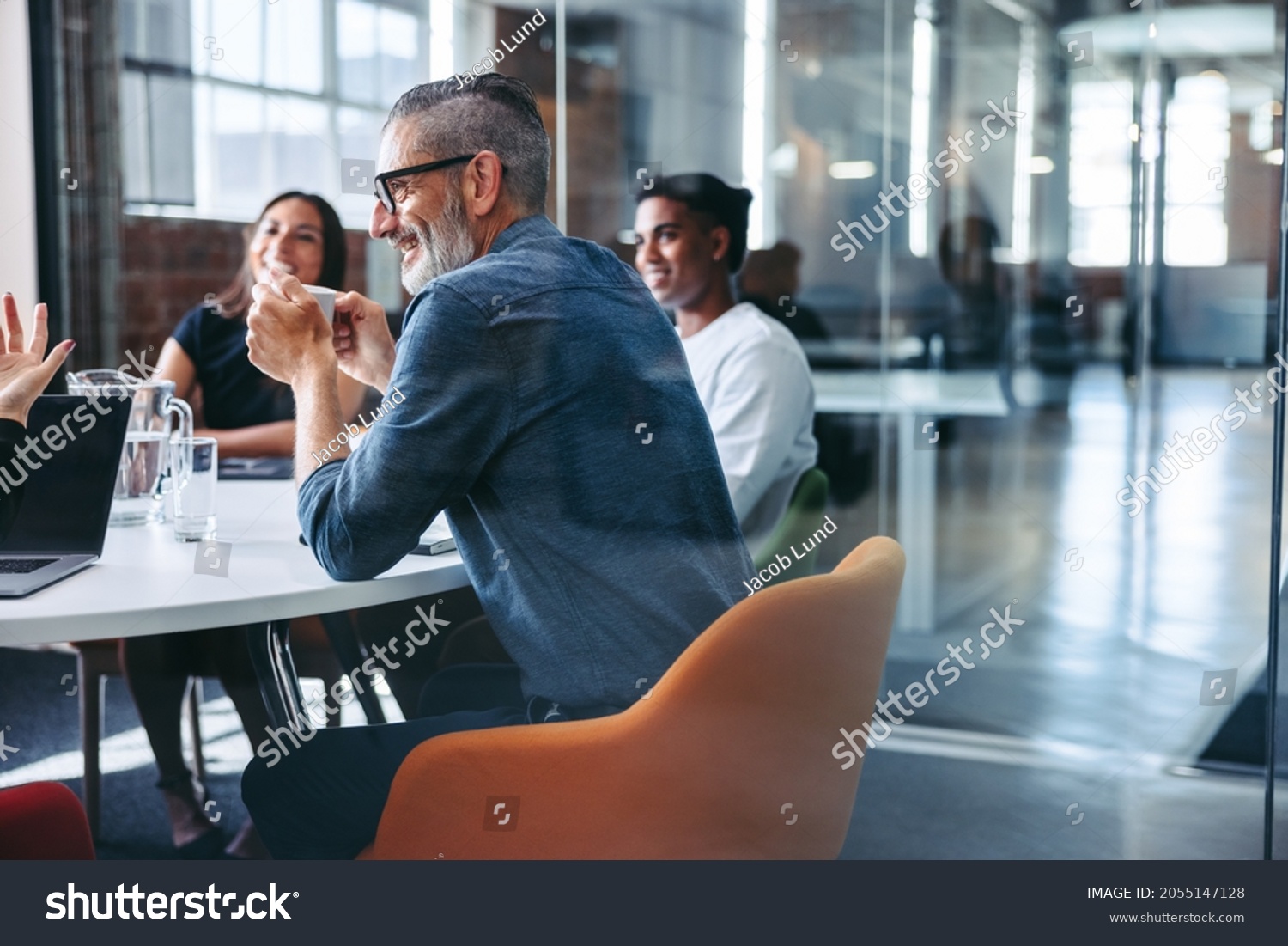 Happy mature businessman attending a meeting with his colleagues in an office. Experienced businessman smiling cheerfully while sitting with his team in meeting room. Businesspeople working together #2055147128