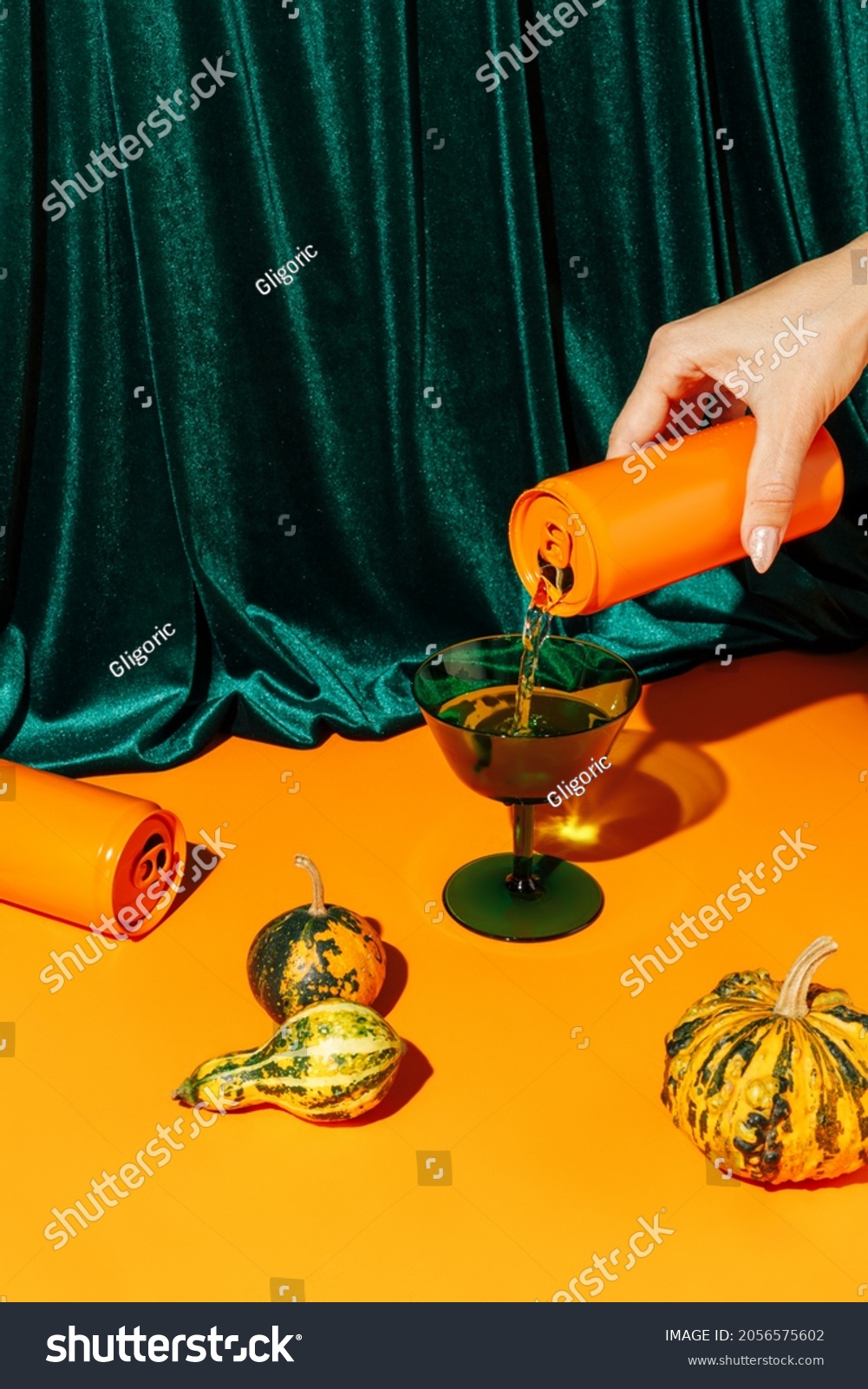 Orange pumpkins and a woman's hand pouring a beverage drink from an orange can against plush velvet curtain background. Creative Halloween and Thanksgiving concept. Contemporary fall still life idea. #2056575602