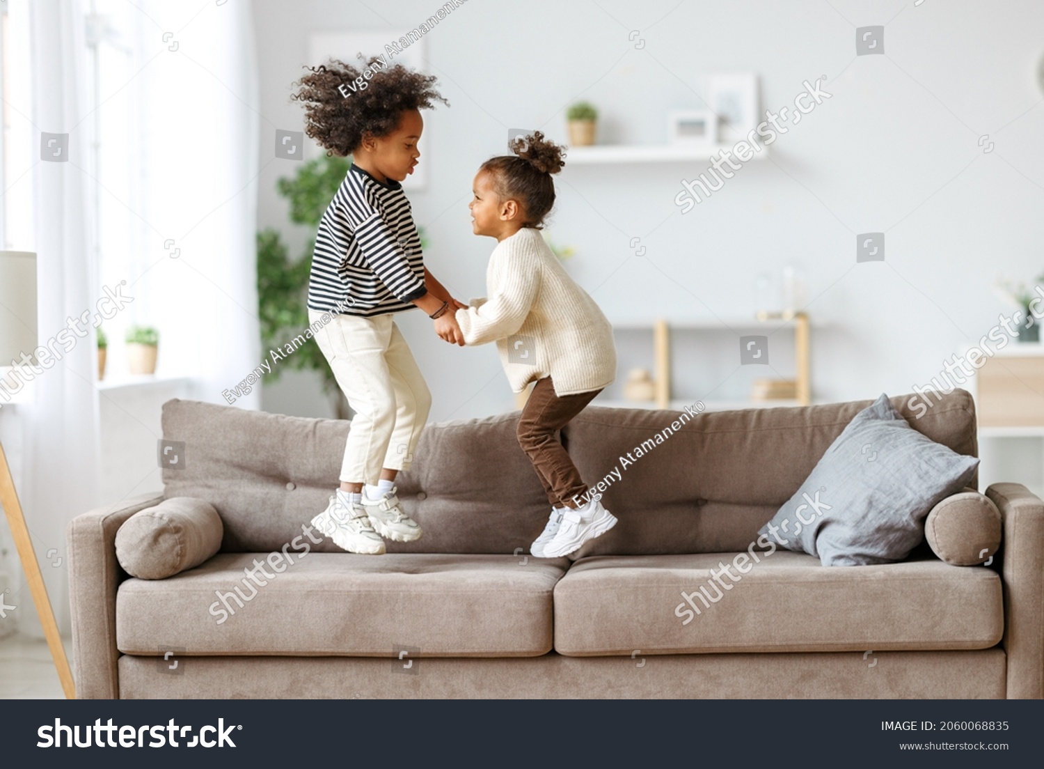 Carefree childhood. Happy energetic african american children jumping on sofa while playing game together at home, small active kids brother and sister having fun in living room #2060068835