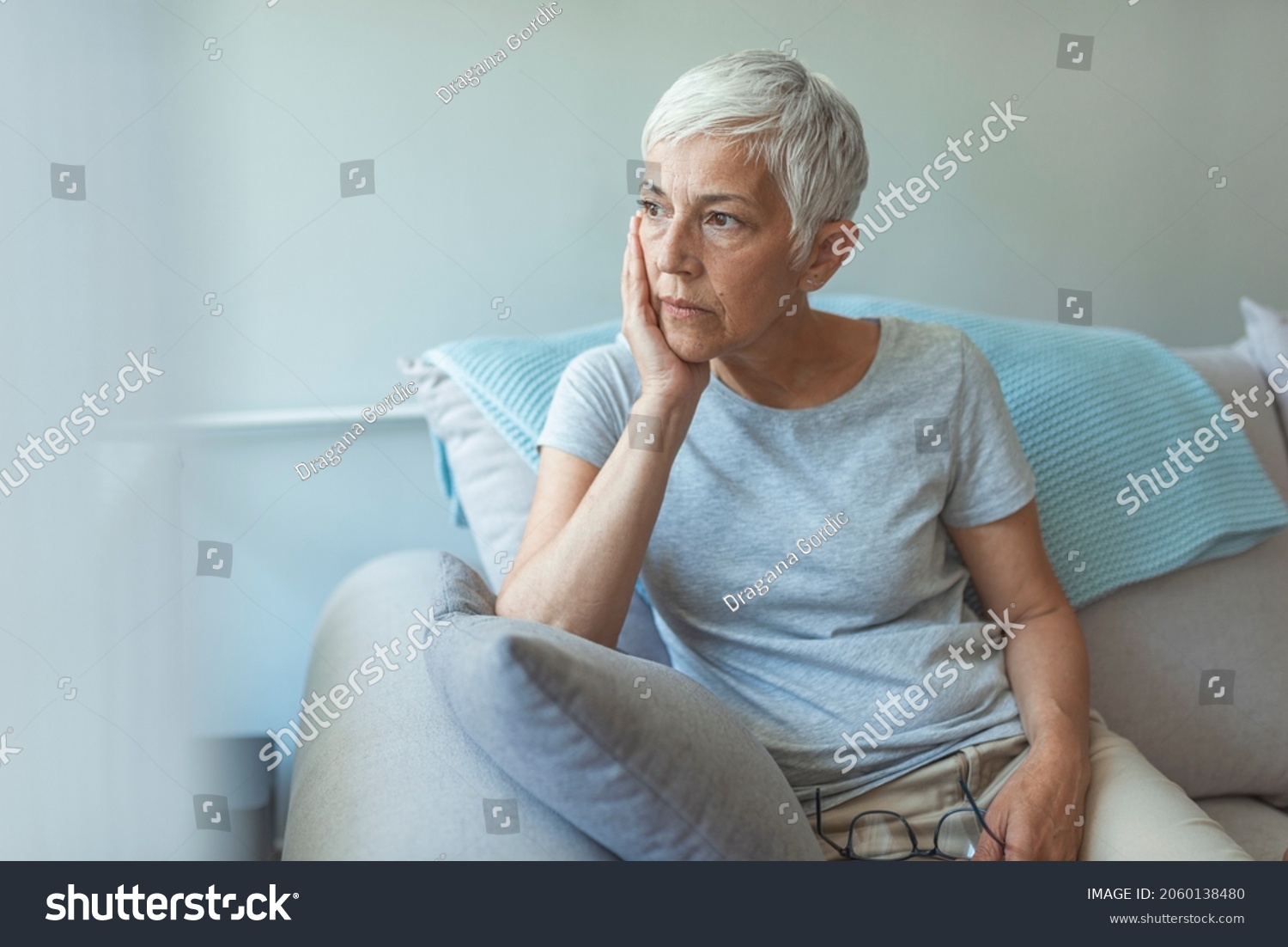 Upset depressed senior gray-haired lady touching temple, sitting on sofa, looking in the window, feeling worried and stressed about the future and thinking about own problems #2060138480