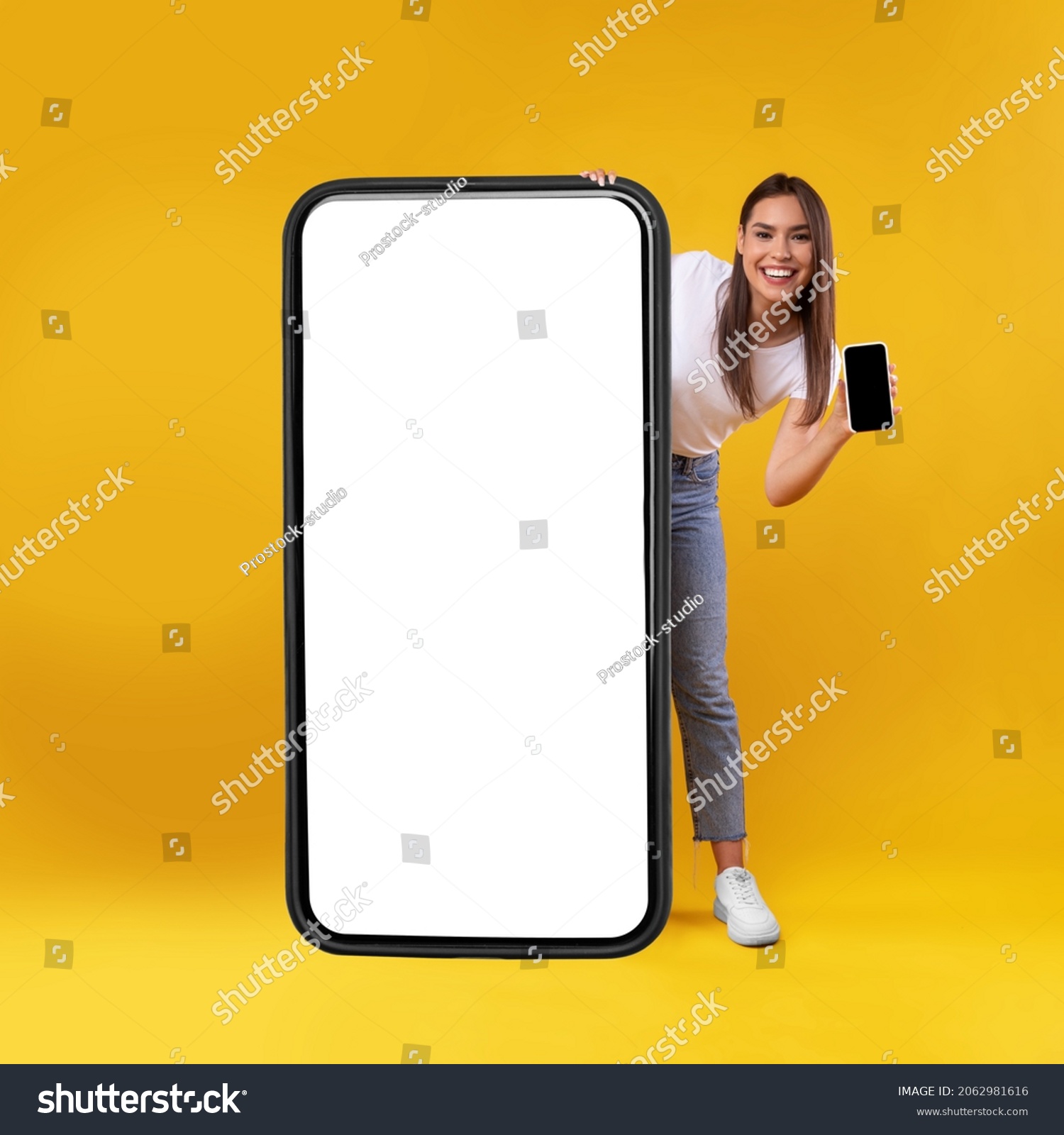 Full Body Length Of Cheerful Woman Peeking Out Standing Behind Big Smartphone With White Blank Screen, Excited Lady Holding Cell Phone Presenting New App Showing Copy Space For Website Design Mock Up #2062981616