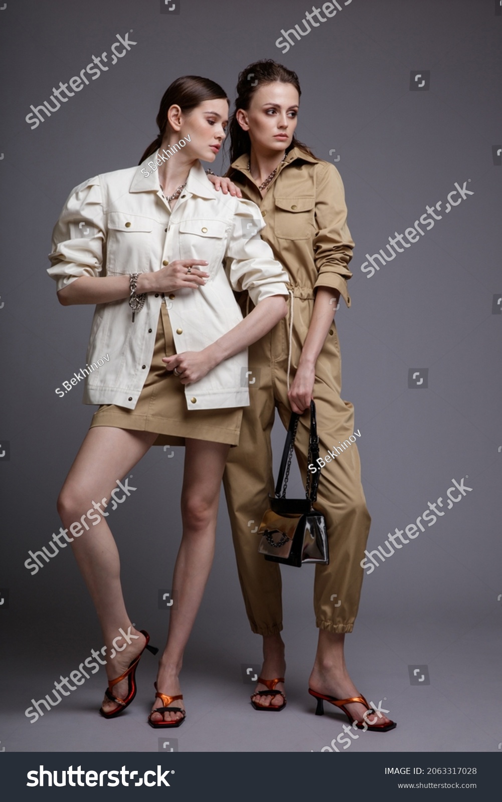 Two high fashion models in white shirt,  beige skirt, jumpsuit, accessories, handbag. Beautiful young women. Studio shot, portrait. Gray background.  Slim figure. Make up, hairstyle #2063317028