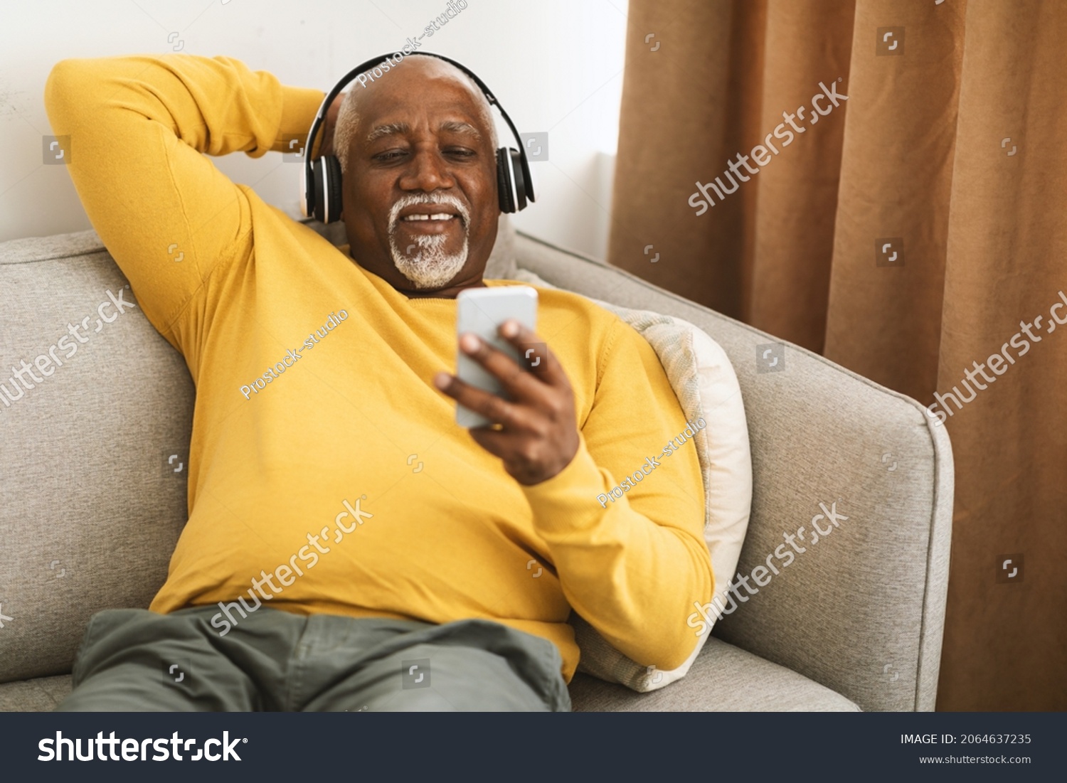 Senior African American Man Listening To Music On Smartphone Sitting On Couch At Home. Male Wearing Headphones Enjoying Favorite Song Or Audiobook On Weekend. People And Gadgets #2064637235