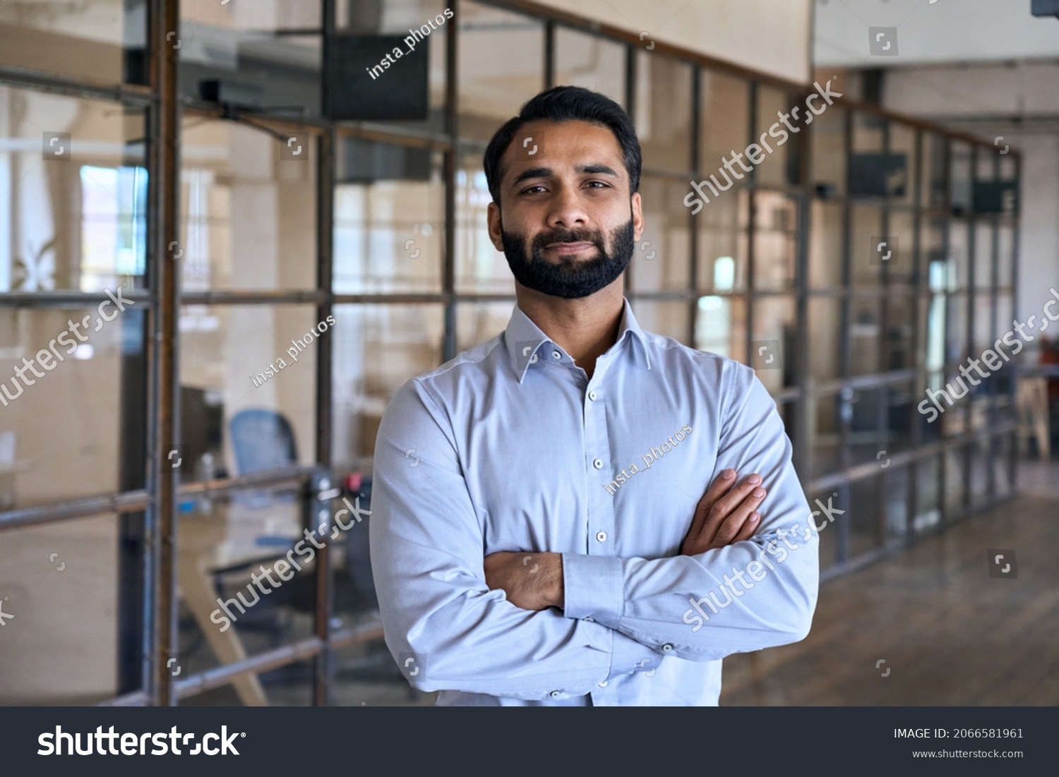 Confident happy successful ceo financial manager. Bearded indian businessman looking at camera standing in modern office with arms crossed. Handsome classy corporation owner. Business portrait. #2066581961