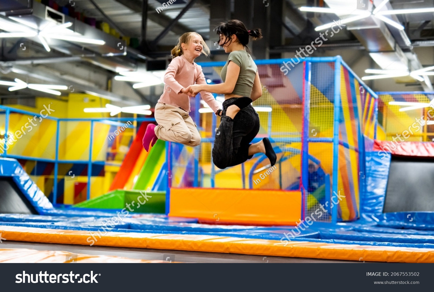 Pretty girls jumping together on colorful trampoline at playground park. Two sisters having fun during active entertaiments indoor #2067553502