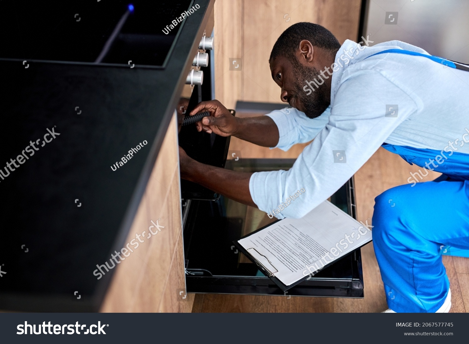 Afro american confident repairman in uniform working, fixing broken oven in kitchen using tools. Black guy in blue workwear is repairing. Repair service concept. Side view portrait #2067577745