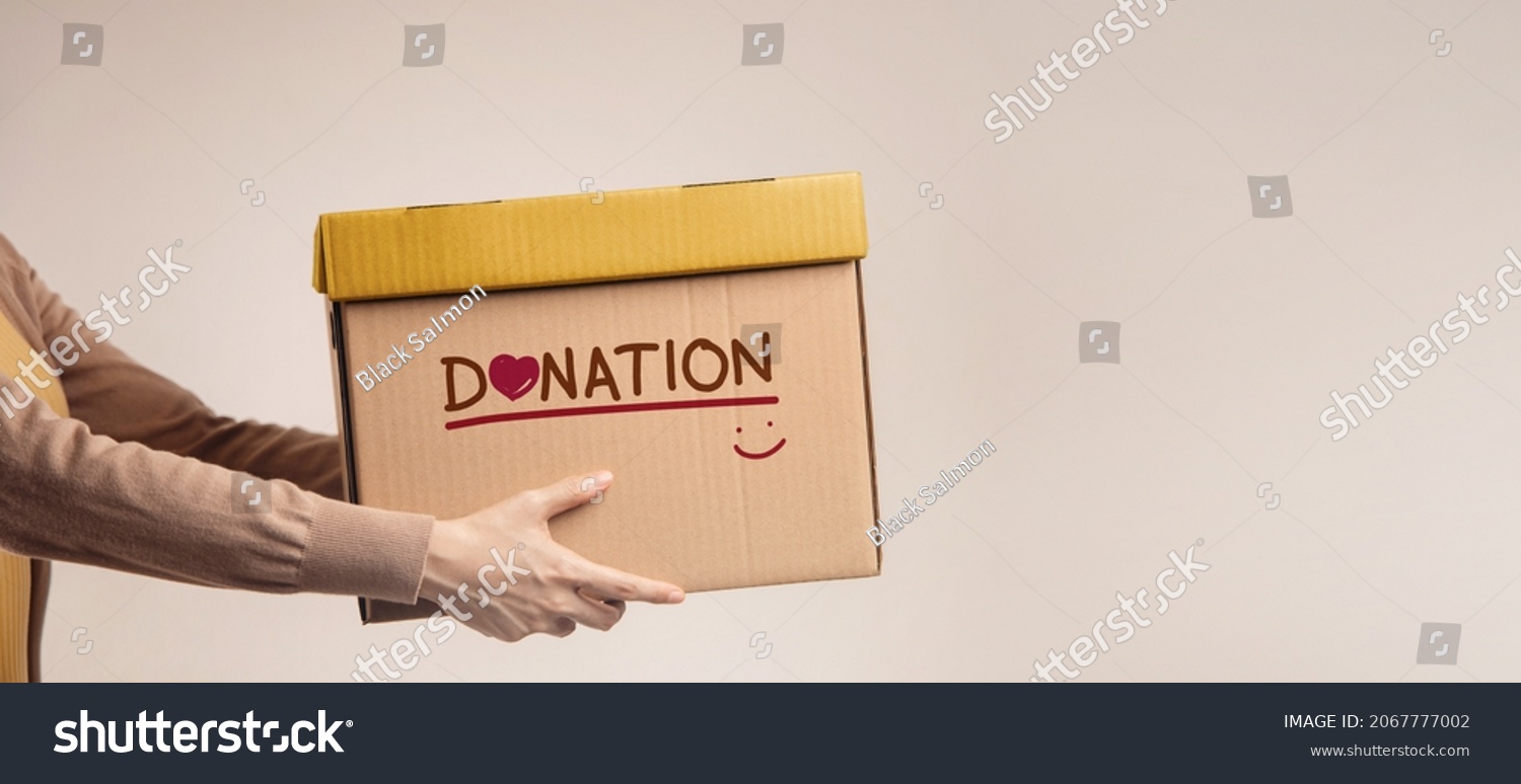 Donation Concept. Woman with Box of Things for Donate with Donation label, Smiling and Heart. Standing against the Walll. Side View #2067777002