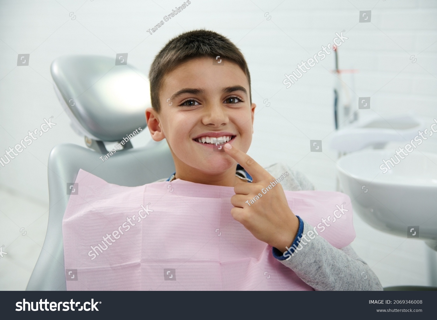 Happy boy holds finger near his mouth, looks at camera, smiles with beautiful toothy smile after receiving dental treatment in dentistry clinic. Oral hygiene, early prevention teeth diseases concept #2069346008