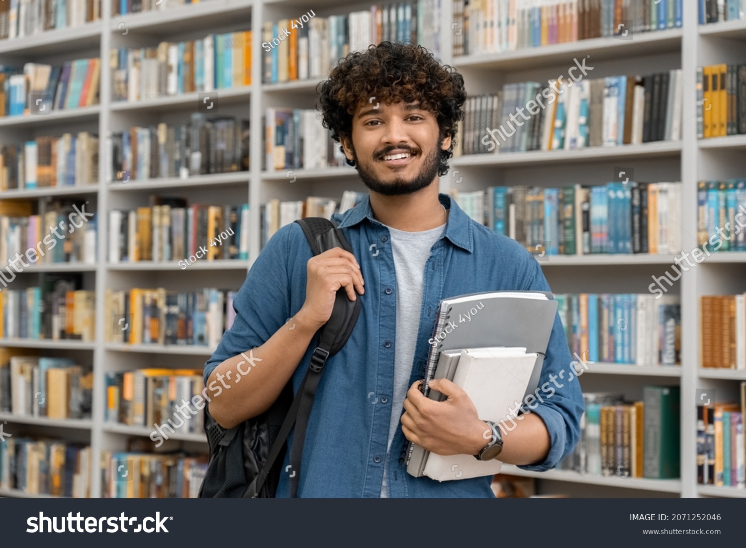 Portrait of cheerful male international Indian student with backpack, learning accessories standing near bookshelves at university library or book store during break between lessons. Education concept #2071252046