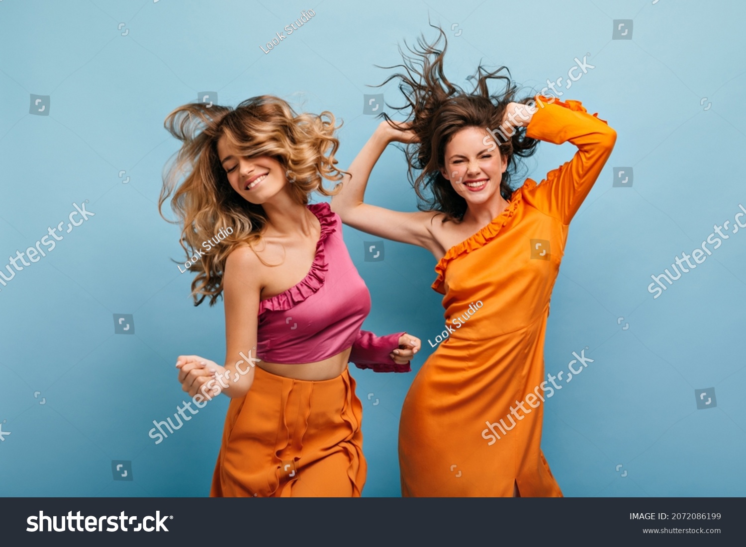Cheerful showy caucasian young women fooling around on isolated blue background. Their wavy, loose hair flies in all directions. Girls are dressed in bright summer outfits. #2072086199