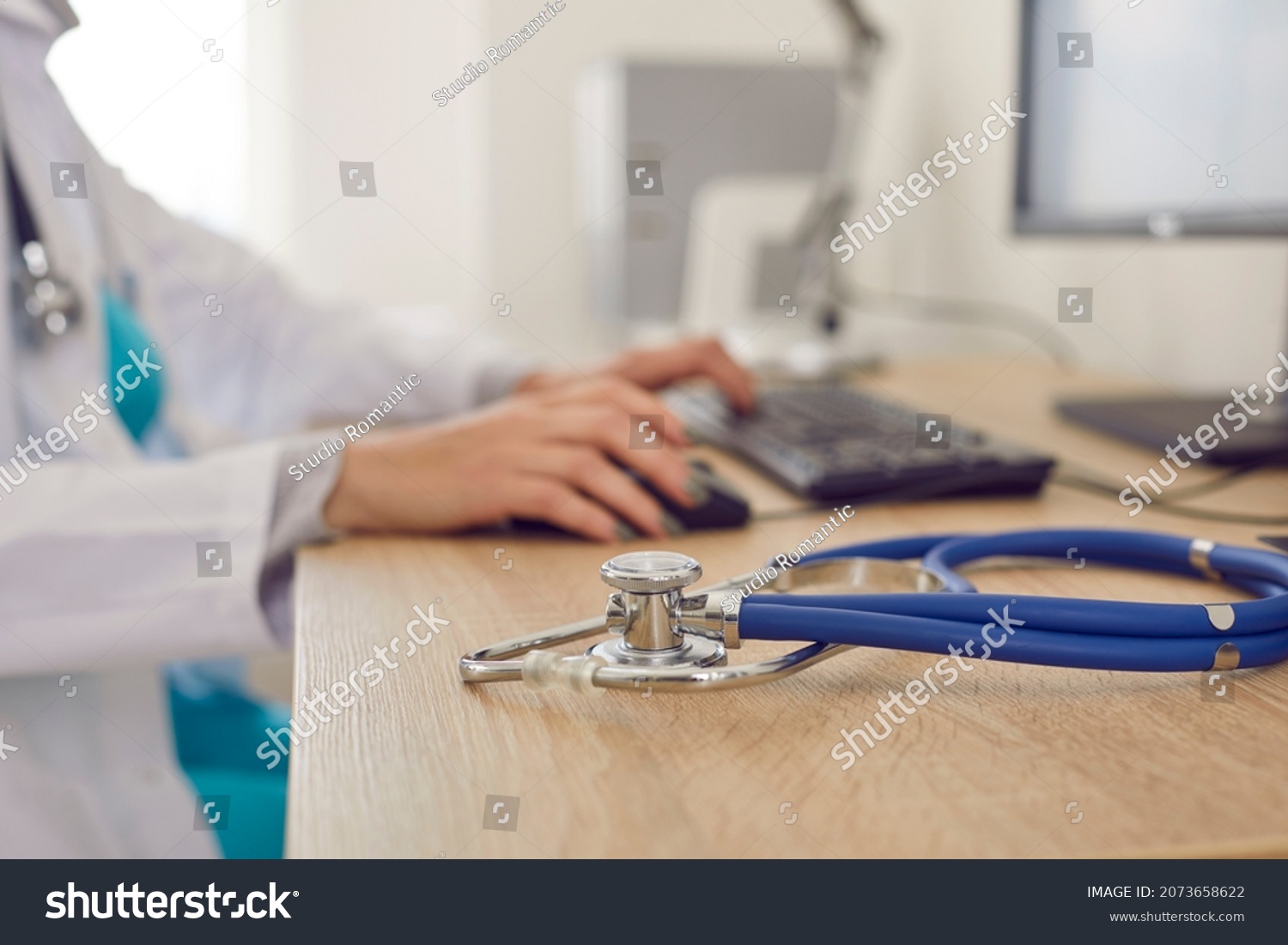 Crop close up of woman doctor sit at desk work on computer in clinic or hospital. Focus on stethoscope phonendocsope on GP table in cabinet. Medical service, healthcare and medicine concept. #2073658622
