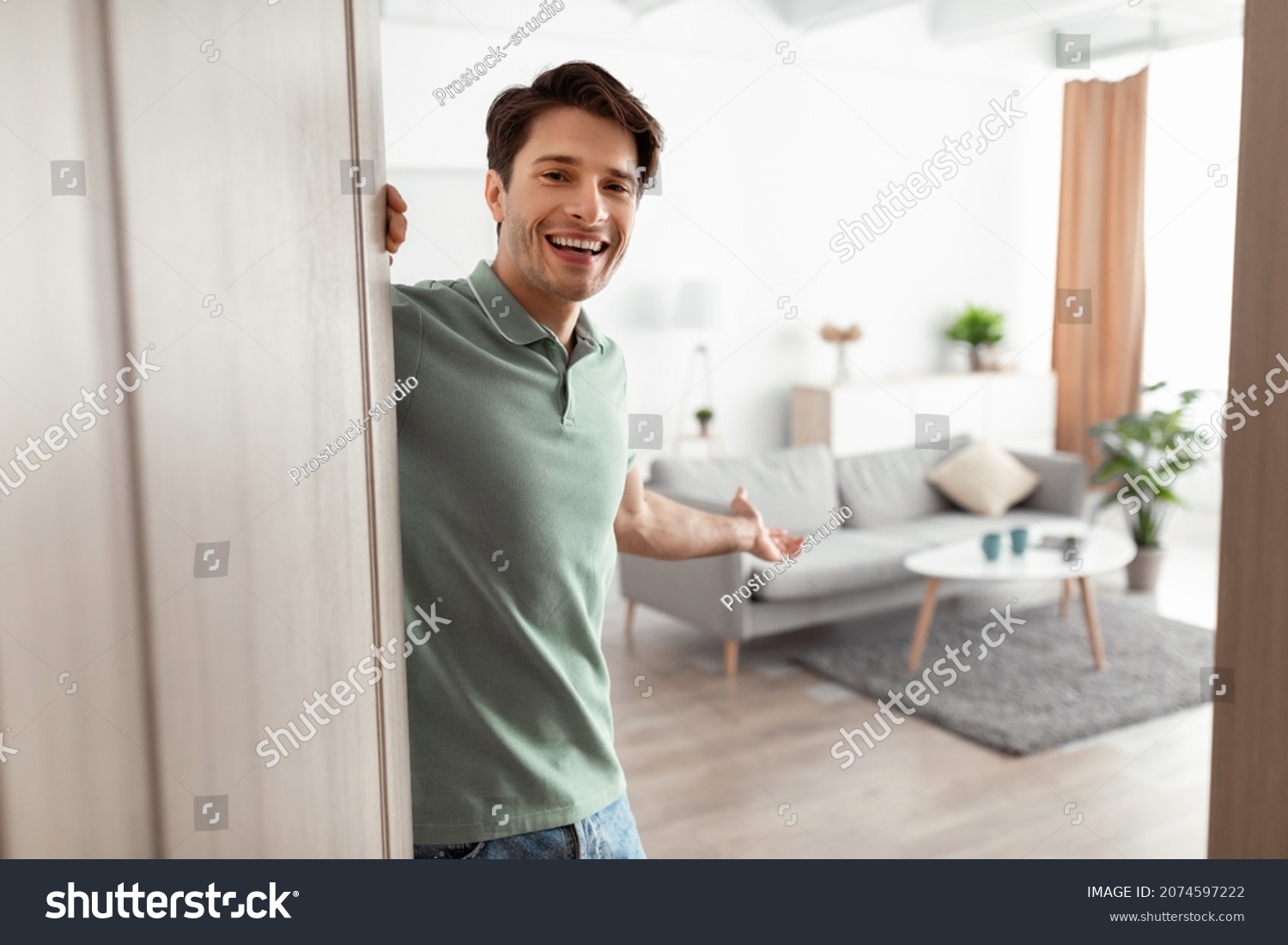 Welcome. Portrait of cheerful man inviting visitor to enter his home, happy young guy standing in doorway of modern apartment, millennial male holding door looking out showing living room with hand #2074597222