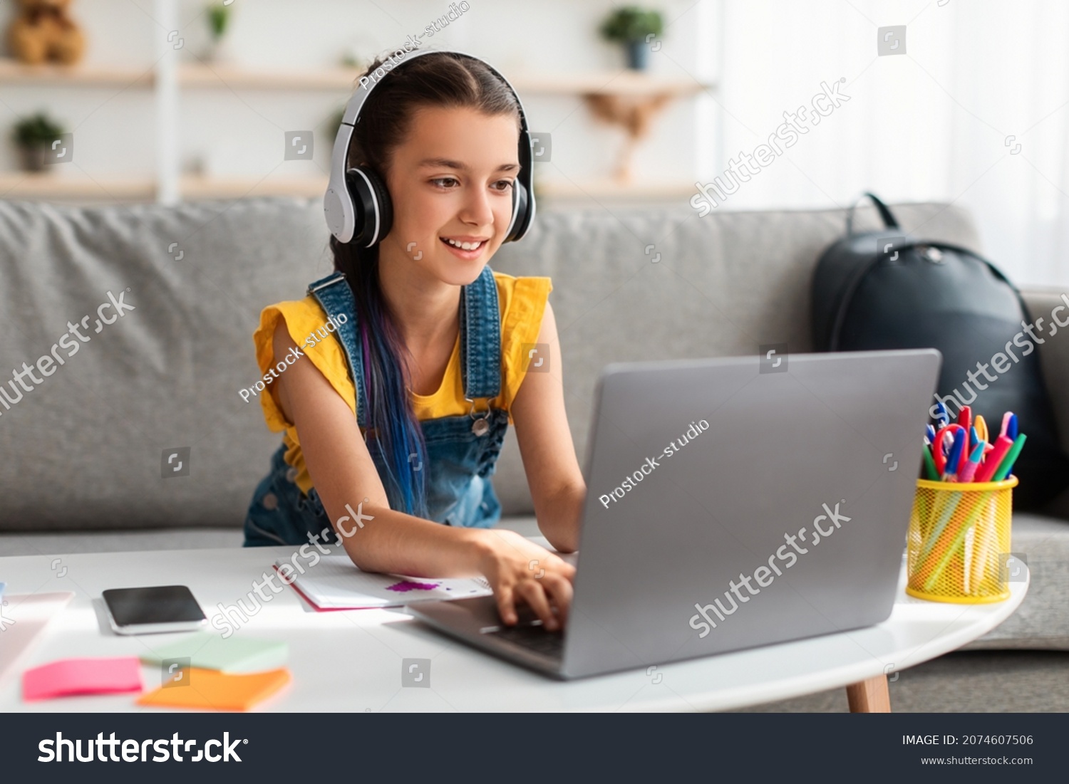 Portrait of little girl in wireless headset using laptop, studying online at home, interested happy student typing on keyboard looking at pc screen, watching webinar, online course, doing homework #2074607506