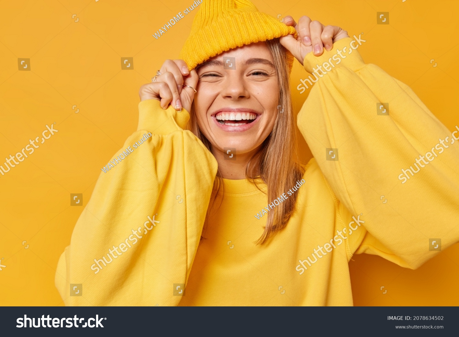 Happy playful young woman smiles broadly shows white teeth keeps hands on hat dressed in casual jumper expresses sincere emotions being in good mood isolated over yellow background. Joy concept #2078634502