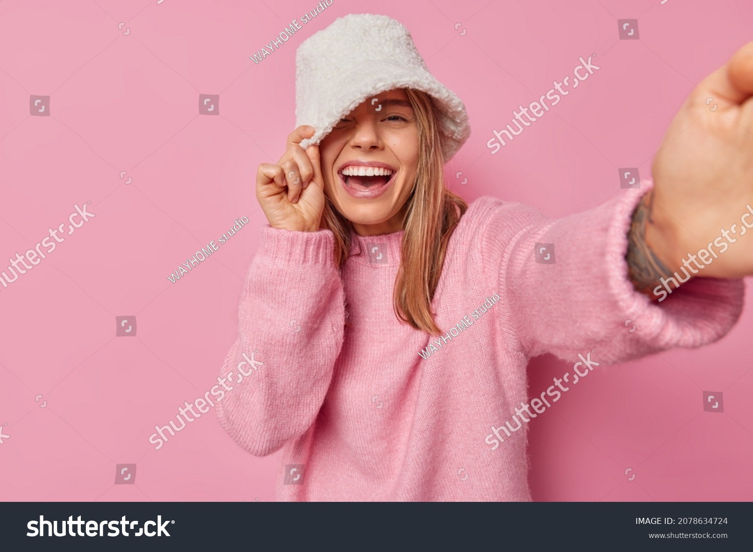 Cheerful optimistic woman wears white panama and jumper has positive playful expression poses for making selfie stretches arm forward camera isolated over pink background takes photo of herself #2078634724