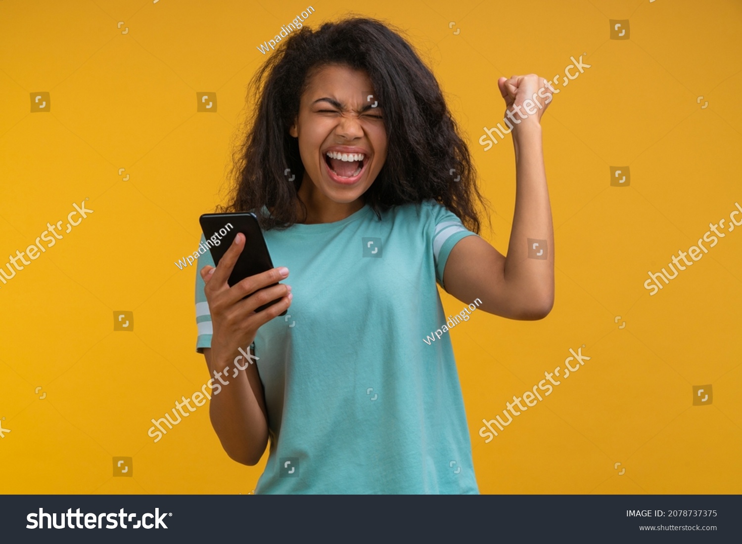 Excited soccer fan girl cheering for favourite team and making winner gesture with her fist being happy about the score. Young woman showing sincere excitement about the victory in online lottery.  #2078737375