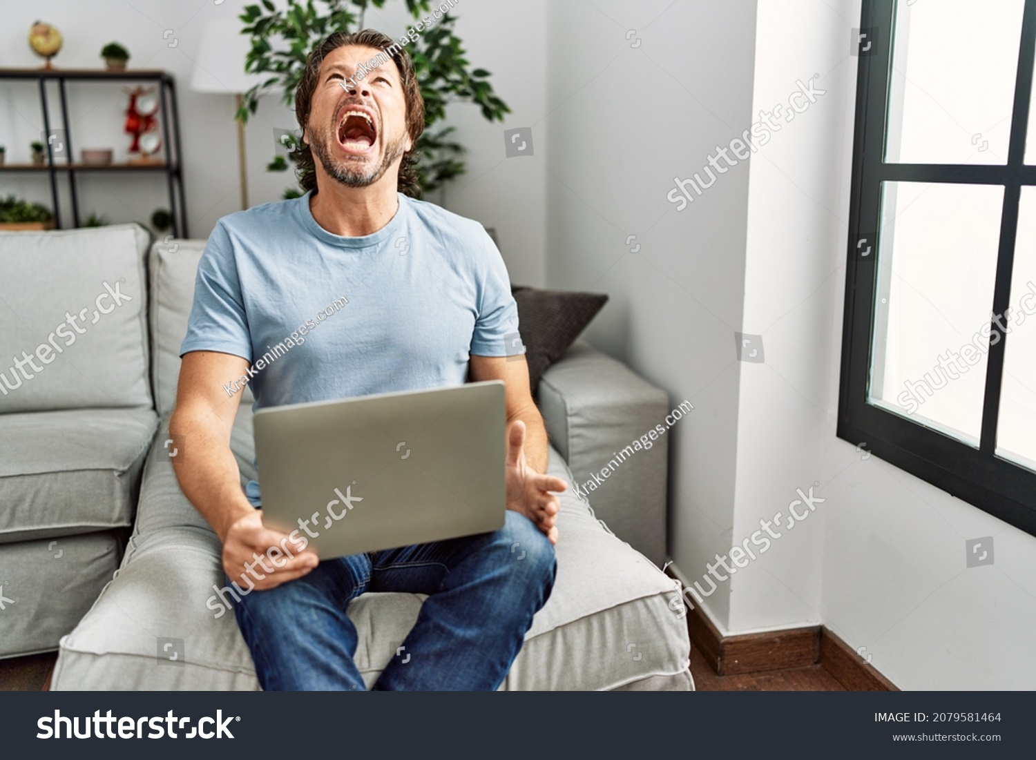 Handsome middle age man using computer laptop on the sofa angry and mad screaming frustrated and furious, shouting with anger. rage and aggressive concept.  #2079581464