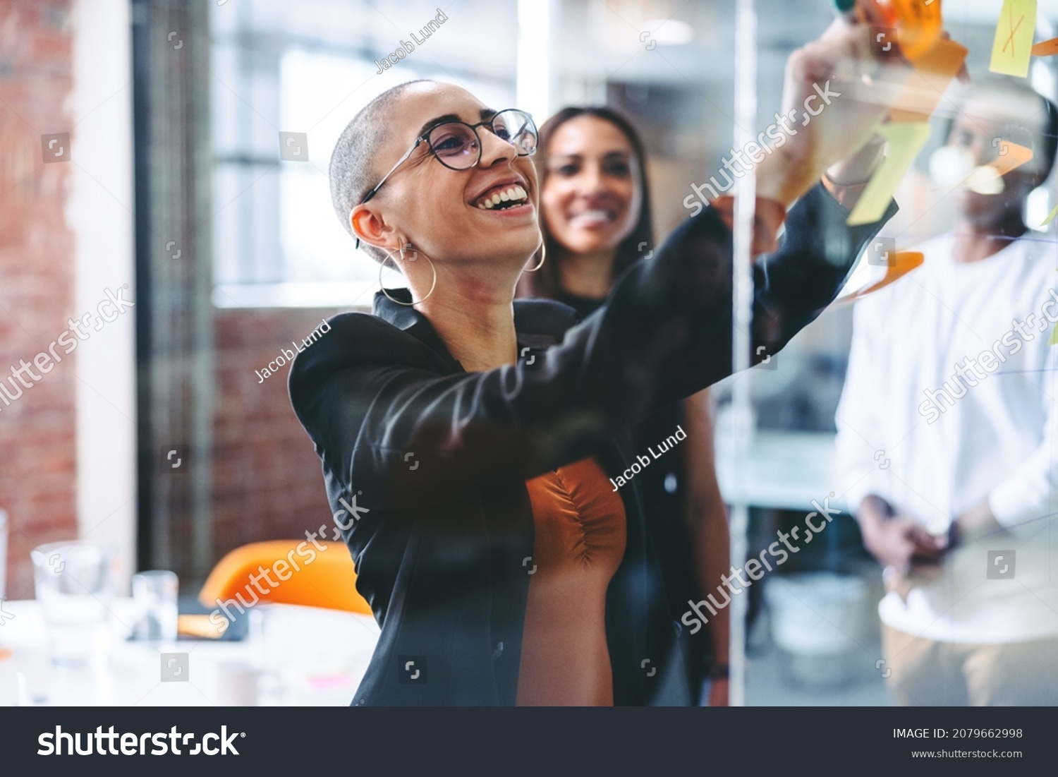 Smiling businesswoman sharing her ideas with her colleagues in a creative workplace. Confident young businesswoman sticking adhesive notes to a glass wall with her team standing in the background. #2079662998