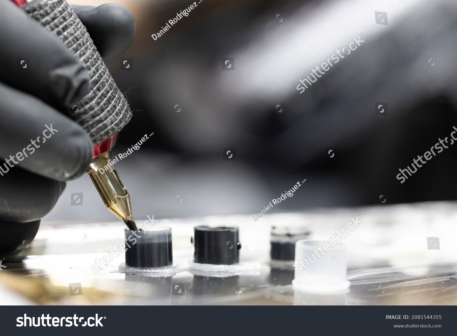Detail of the tattooist's hand with black glove holding the tattoo machine while he loads ink into the needle while performing a tattoo. Image in horizontal and colour. #2081544355