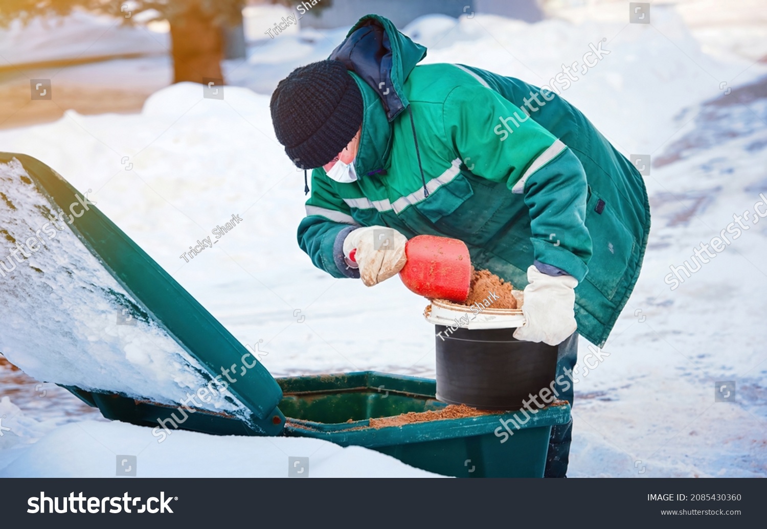 Municipal worker in uniform scoop sand from grit bin, spreading deicing chemicals on slippery sidewalk. Sanding street, prevent injury and slipping accident #2085430360