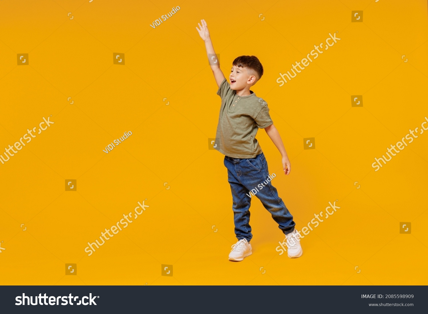 Full body side view little small smiling happy boy 6-7 years old wear green t-shirt walk go waving hand isolated on plain yellow background studio portrait. Mother's Day love family lifestyle concept #2085598909