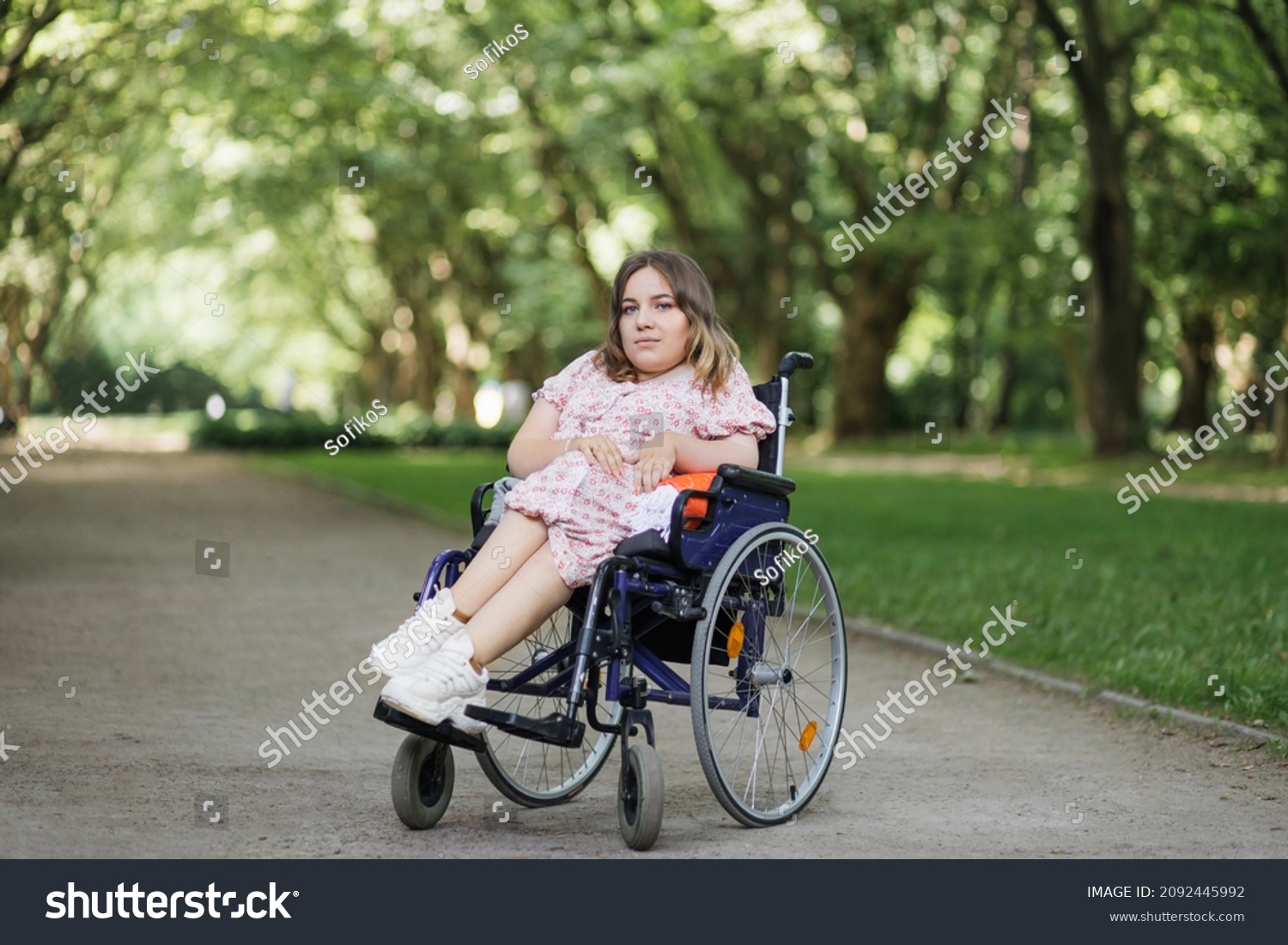 Portrait of pleasant young woman with spinal muscular atrophy smiling on camera among green summer park. Female person who using wheelchair. Concept of people with disability. #2092445992