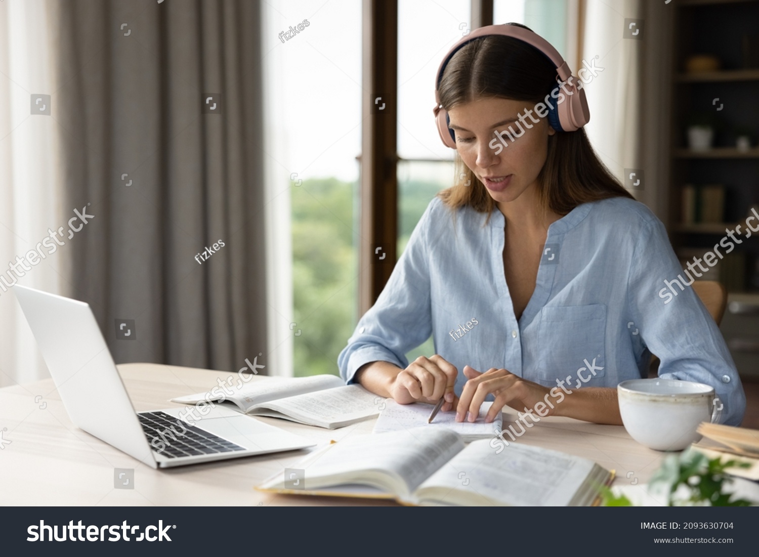 Focused engaged student girl in big headphones studying foreign language, listening audio lesson at laptop, reading notes out loud, doing exercises from open book, school, college homework task #2093630704