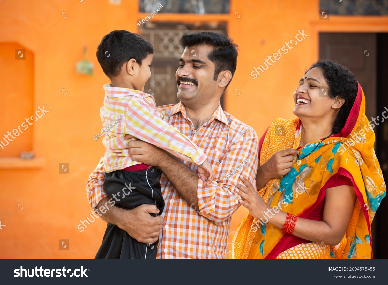 Young happy indian parents holding cute little child having fun and laughing while standing outdoor at village house. Mustache man wearing kurta and woman wear sari. rural india concept. #2094575455