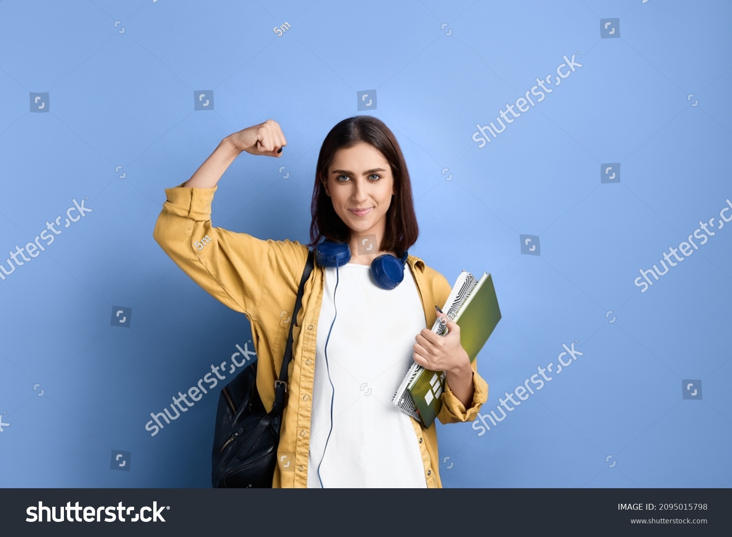 Good luck. Strong smiling student girl shows muscles, passed a milestone, confident in her knowledge, holding books, wearing yellow shirt, white t-shirt, black bag and headphones over neck #2095015798