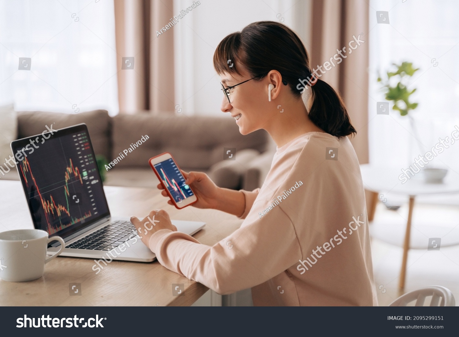 Smiling business woman trader analyst looking at laptop monitor, holding smartphone, wearing earphones. Investor broker analyzing indexes, trading online investment data on stock market graph at home #2095299151