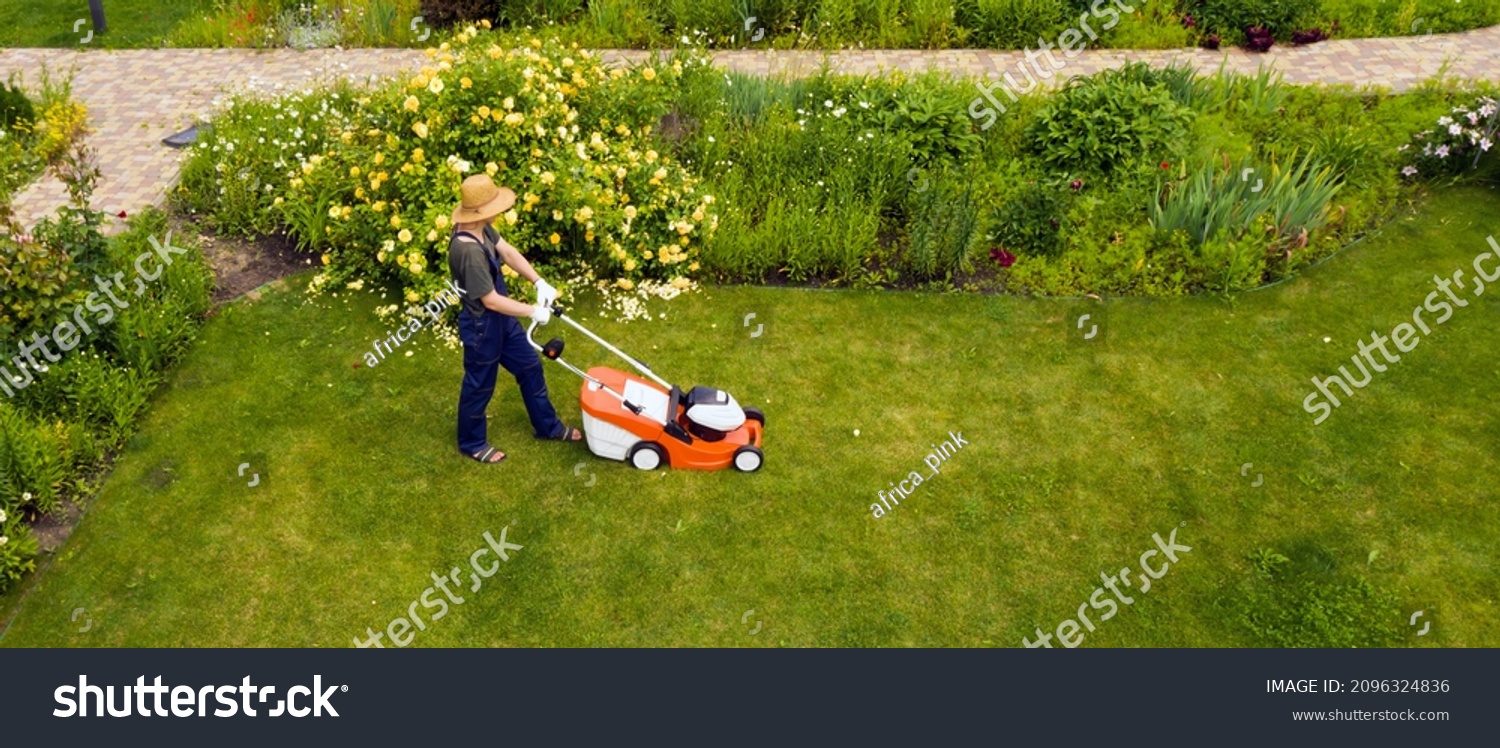 A young man is mowing a lawn with a lawn mower in his beautiful green floral summer garden. A professional gardener with a lawnmower cares for the grass, view from above #2096324836