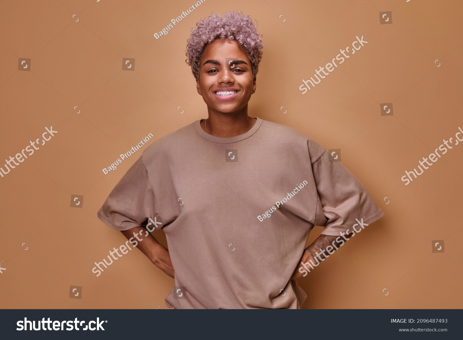 Young attractive ethnic African American woman curly hairstyle smiles broadly and looks at camera holds hands on belt posing standing in beige oversized t-shirt on brown studio background #2096487493