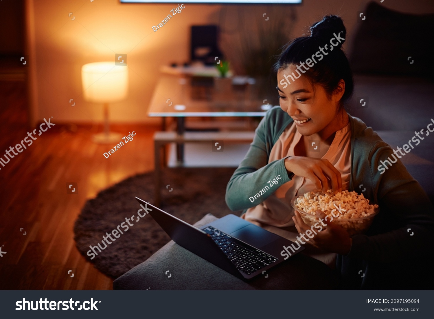 Happy Asian woman eating popcorn while watching her favorite series online on laptop in the evening at home. #2097195094