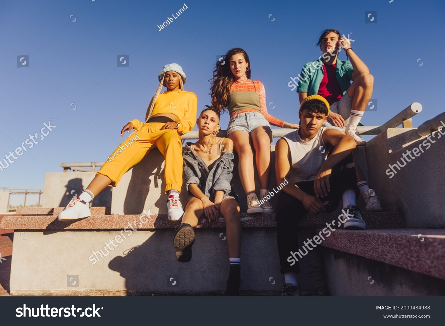 Group of friends sitting together outdoors in the sun. Multiethnic youngsters spending quality time together in the city. Group of generation z friends chilling outdoors. #2099484988