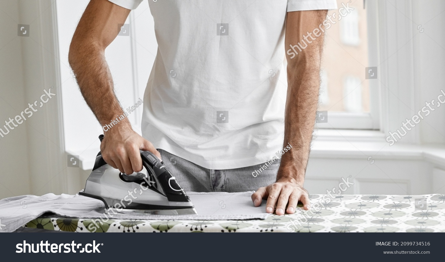 Hard-working bearded man dressed in white t shirt, irons shirt on ironing board, takes care of clothes, does household duties, poses against window background. Housework and masculinity concept #2099734516