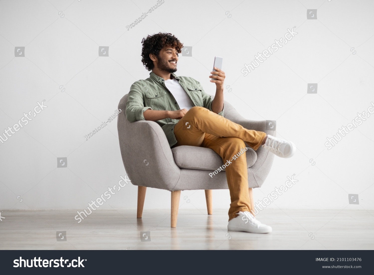 Cheerful curly young indian man chatting with girlfriend while relaxing in armchair at home, using modern mobile phone, checking social media, using mobile app, panorama with copy space #2101103476