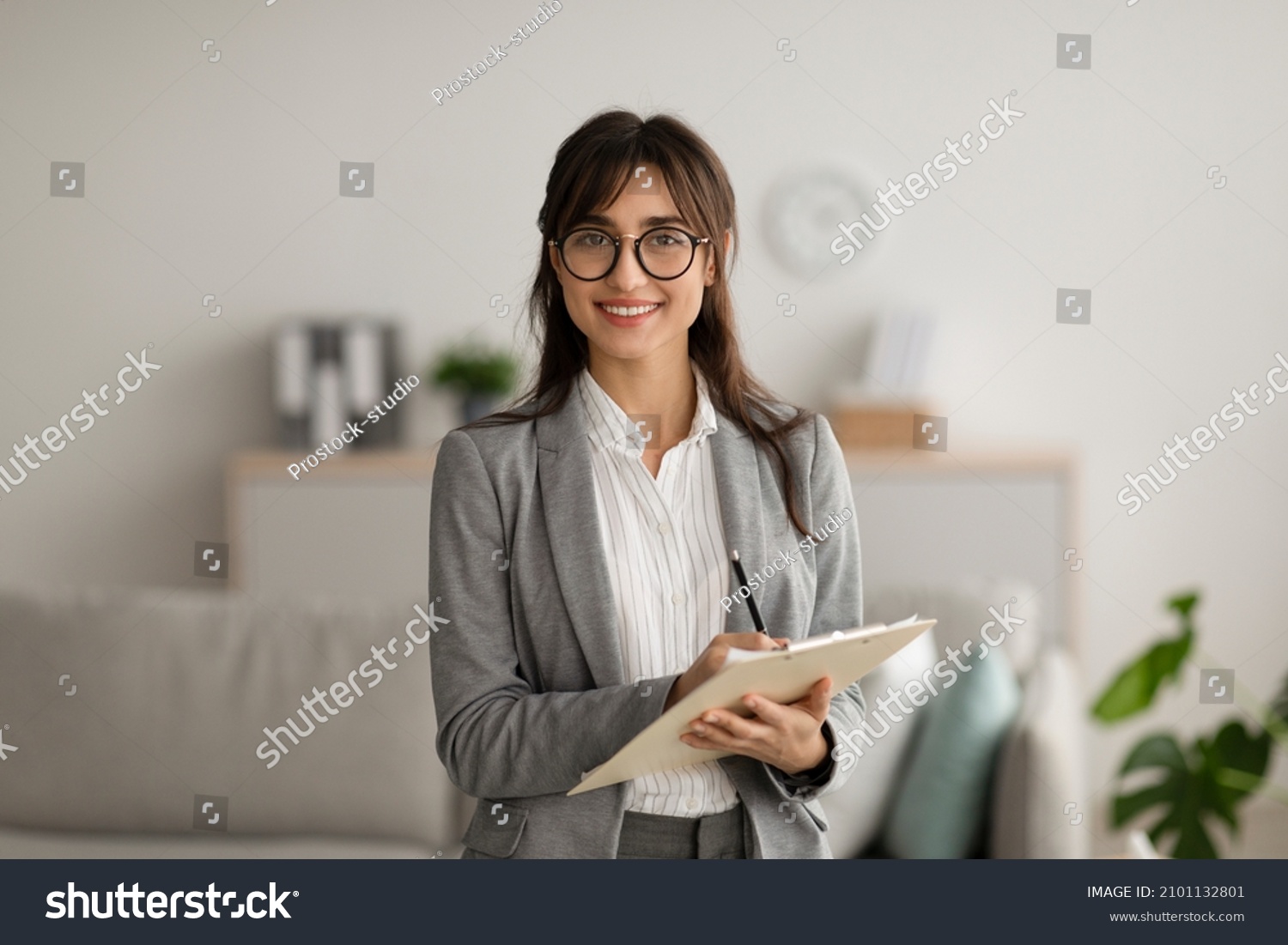 Happy professional middle eastern female psychologist writing in clipboard, looking and smiling at camera, working in modern office. Psychotherapy services, mental health professional concept #2101132801