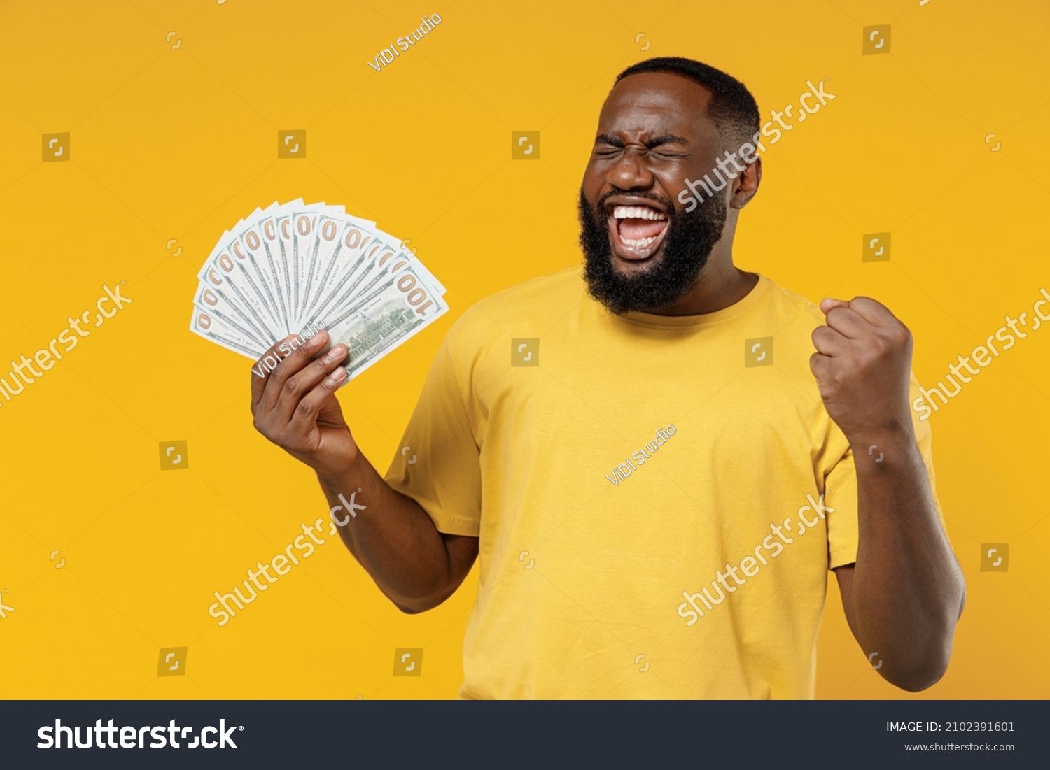 Young smiling happy rich black man 20s wearing bright casual t-shirt holding fan of cash money in dollar banknotes do winner gesture isolated on plain yellow color background. People lifestyle concept #2102391601