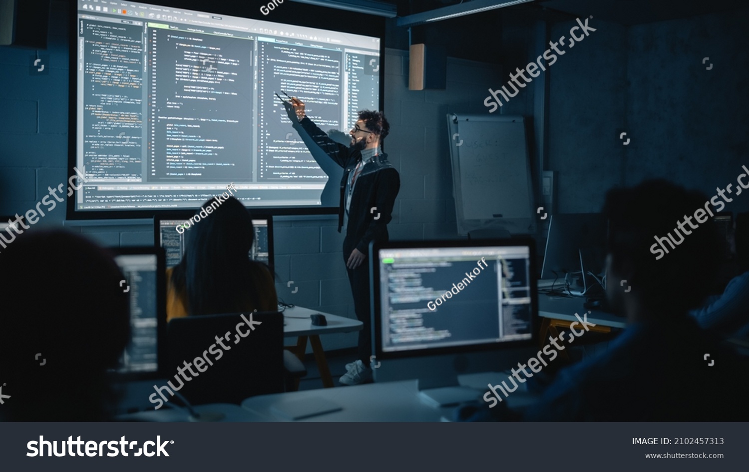Teacher Giving Computer Science Lecture to Diverse Multiethnic Group of Female and Male Students in Dark College Room. Projecting Slideshow with Programming Code. Explaining Information Technology. #2102457313
