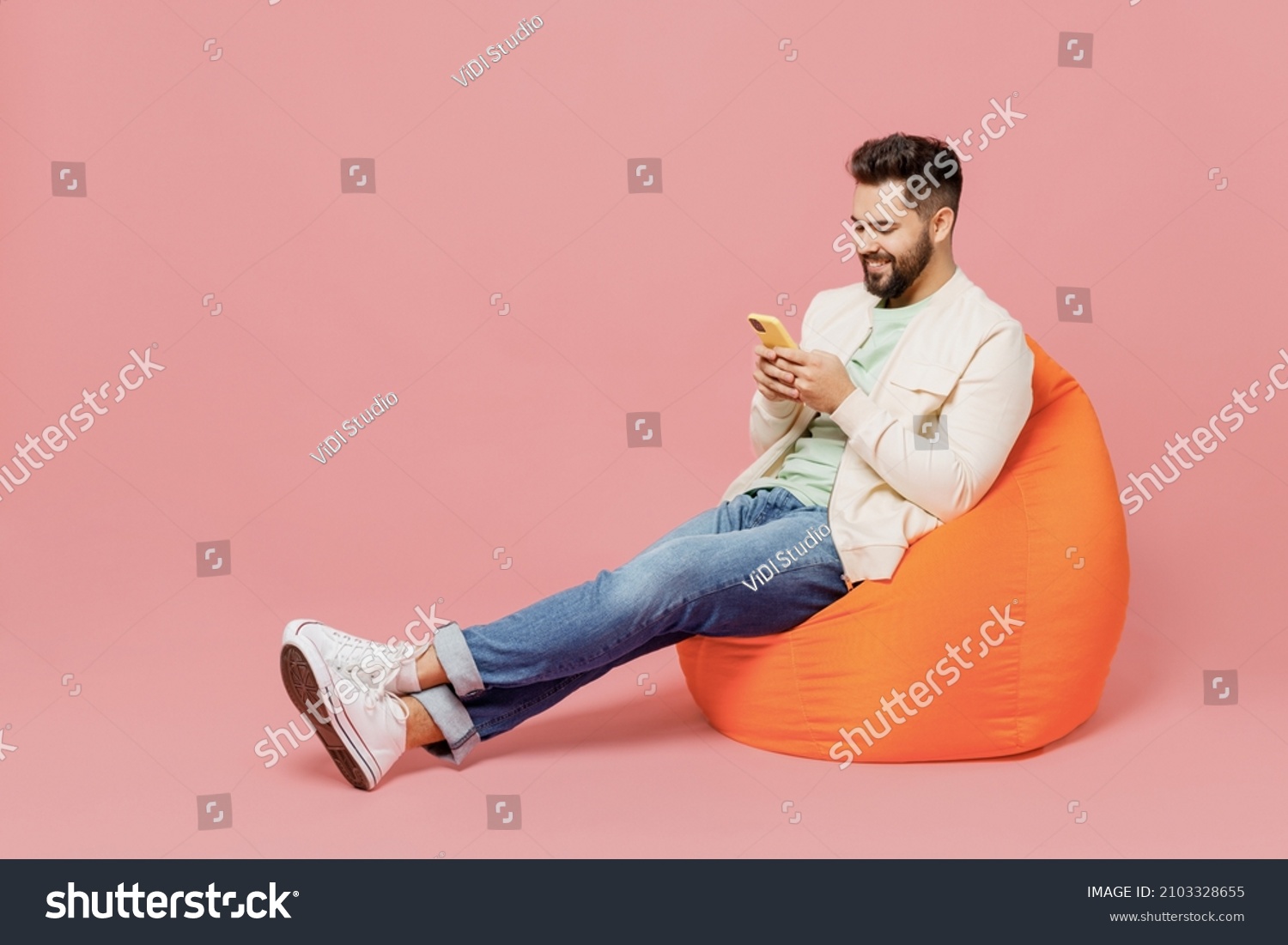 Full body young smiling cheerful happy man 20s in trendy jacket shirt sit in bag chair hold in hand use mobile cell phone isolated on plain pastel light pink background studio People lifestyle concept #2103328655