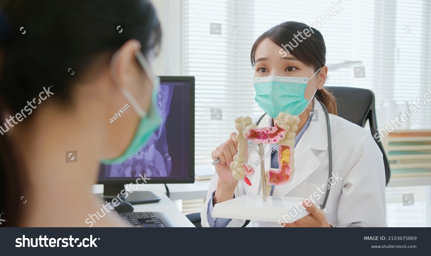 asian woman wears face mask to prevent COVID19 has colorectal cancer diagnosis in hospital - female doctor showing colonoscopy results and xray to patient on computer and explain by anatomical model #2103675869