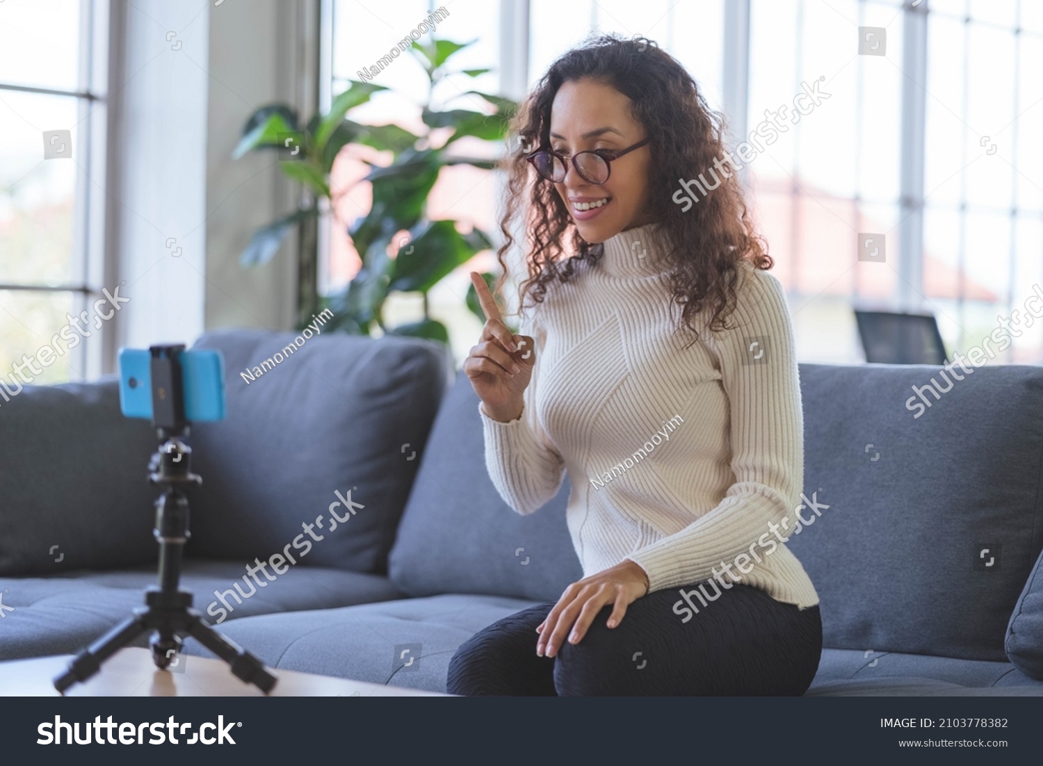 Young African American women wearing eyeglasses live streaming training on the sofa through the application from smartphone to social media during the coronavirus outbreak. #2103778382