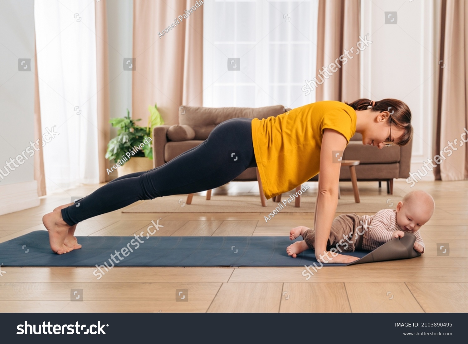 Beautiful yogi mom smiling at her baby while doing push up on exercising mat at home. Happy mom working out with her son. Young mom looking at her little baby during her post-natal fitness routine #2103890495