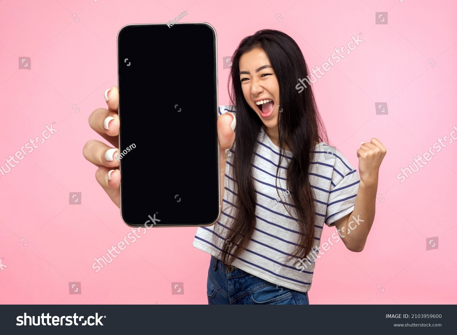 Portrait of euphoric happy joyful girl with long hair screaming with happiness showing mobile advertisement mockup area and celebrating her victory. indoor studio shot isolated on pink background #2103959600
